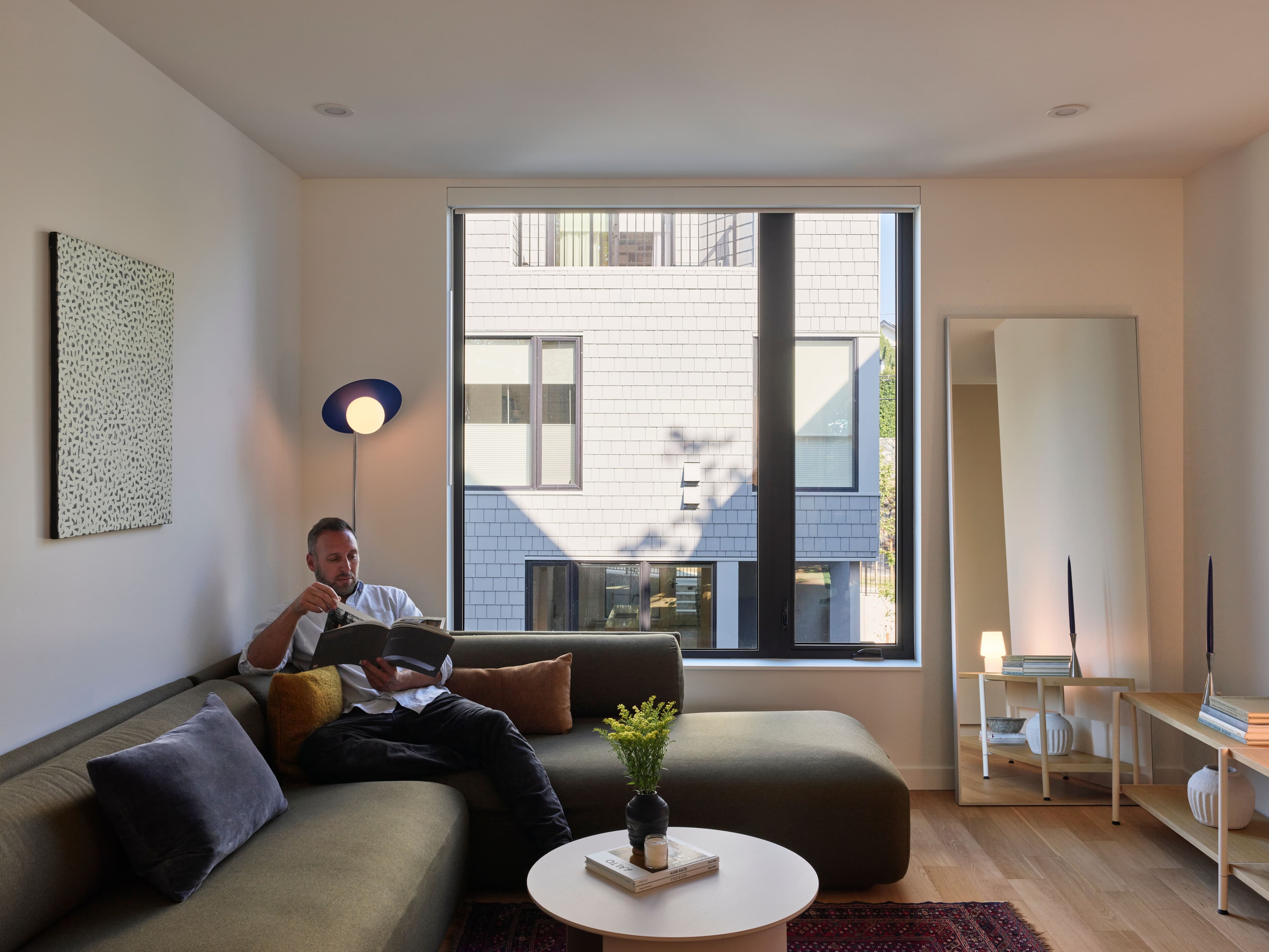 A resident reads on an olive green sectional in the living room of a Wisteria Row townhome in Victoria BC, with a large window and wood floors.