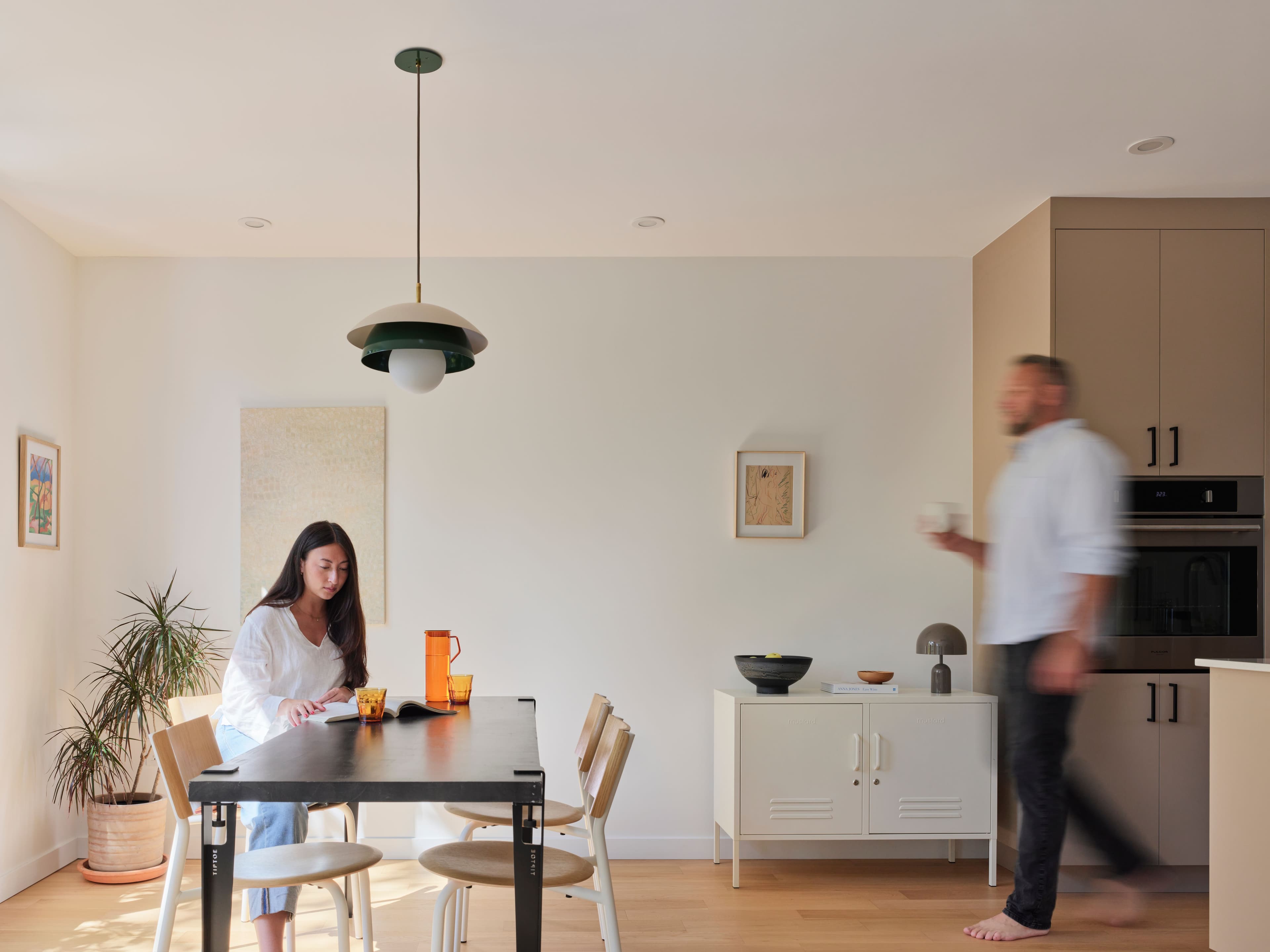 Two residents share a morning in the dining area of a Wisteria Row townhome, one seated at the table and one walking past the kitchen with a mug.