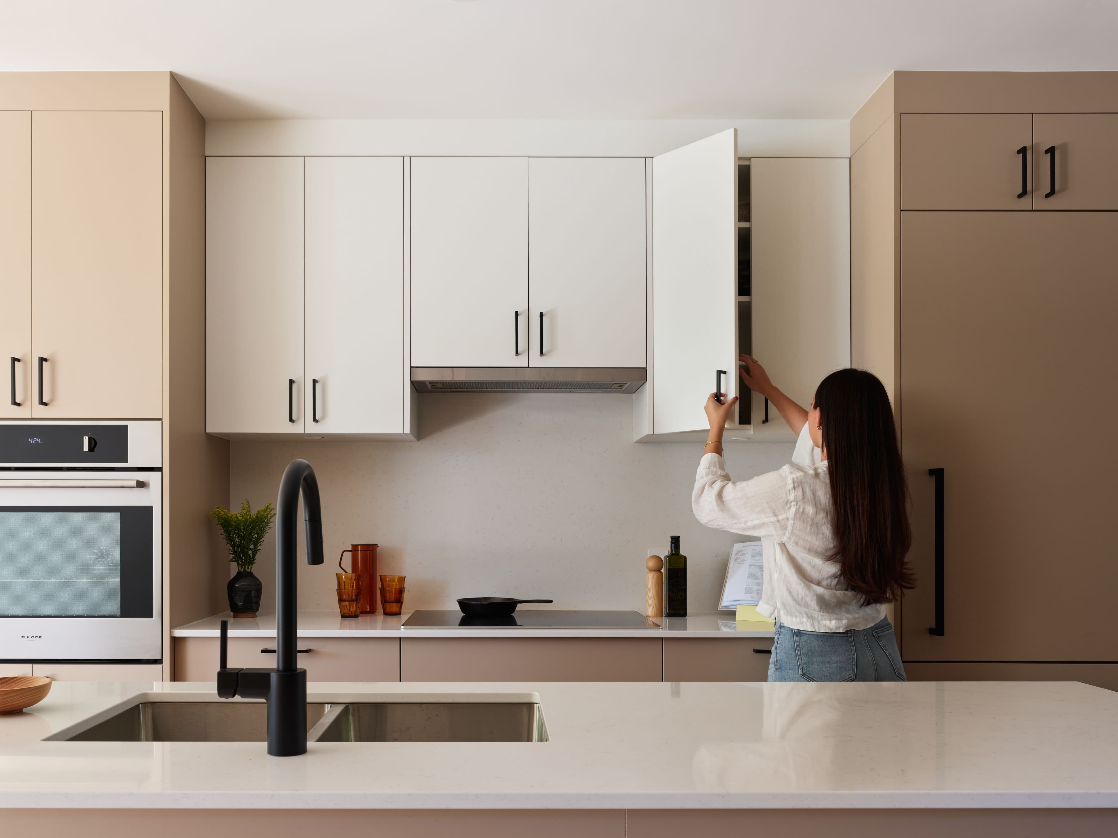 A resident reaches into an upper cabinet in the kitchen of a Wisteria Row townhome, with a quartz island, matte black faucet and wall oven.